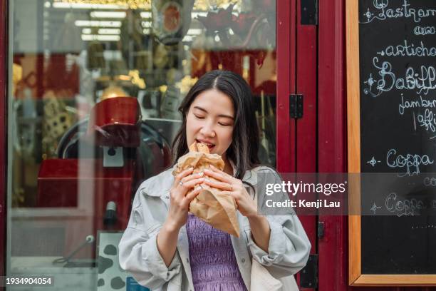 joyful young asian woman enjoying local food in front of a food shop while visiting paris. concept of holiday, travel, fast food, eating out. - nice france stock pictures, royalty-free photos & images