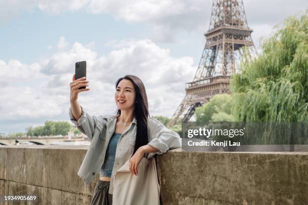 cheerful young asian woman taking selfie by using mobile phone against eiffel tower while having a relaxing time enjoying the sunny day under the clear blue sky in the city. lifestyle and technology. - day to the liberation of paris photos et images de collection