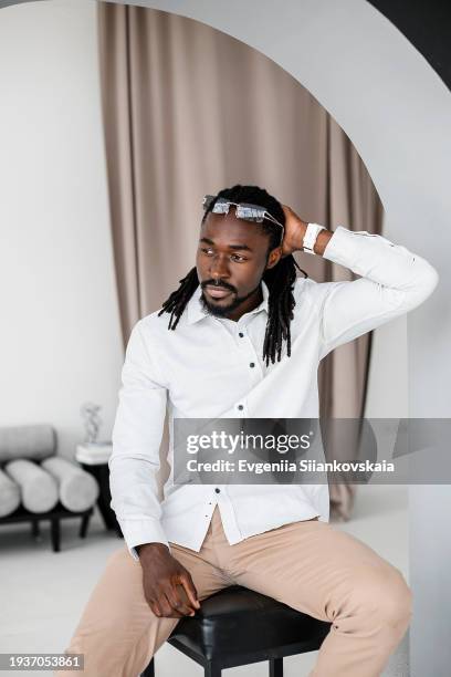 full body portrait of pensive young african man sitting on the chair at living room. - portrait of pensive young businessman wearing glasses stock-fotos und bilder