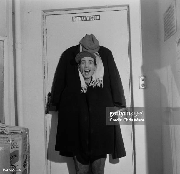 British comedian, actor and singer Norman Wisdom, his body hidden behind the coat hanging from the door to his dressing room, backstage at the London...