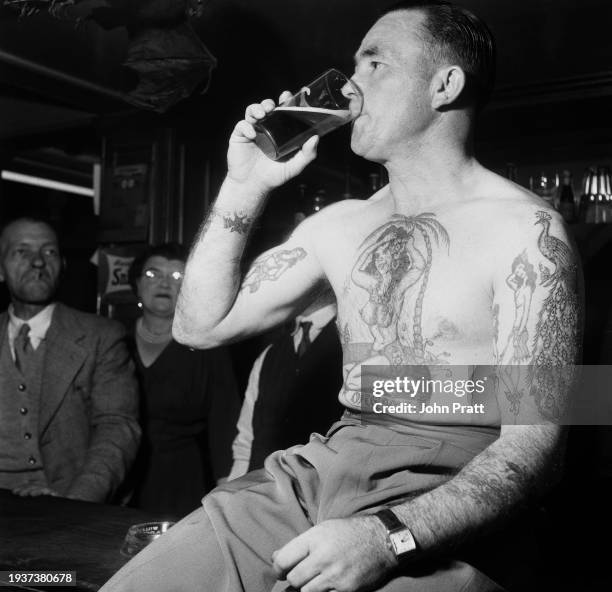 Tattooed man sits shirtless as he drinks from a pint glass, a tattoo of a semi-naked woman on a desert island, captioned 'Dreams' on his chest, at a...