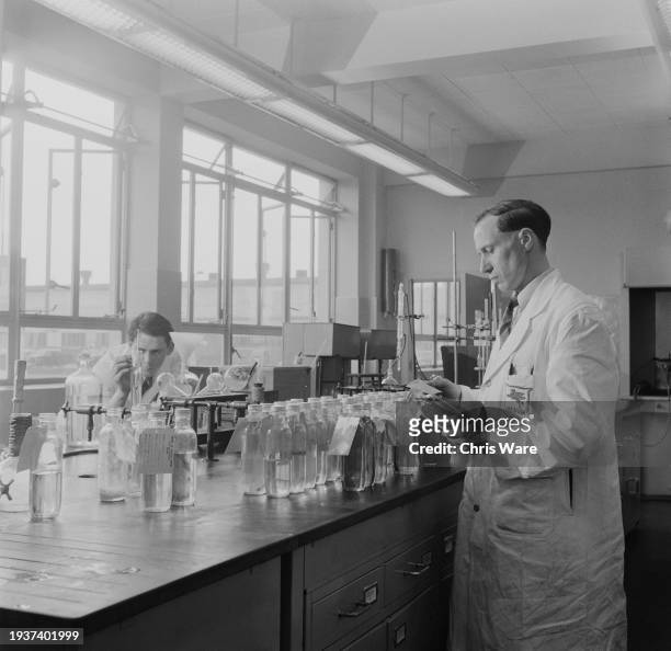 Lab technicians, one wearing a 'Vacuum Oil Company' lab coat examining samples of 'mobile gas', while the other determines the Specific Gravity of a...
