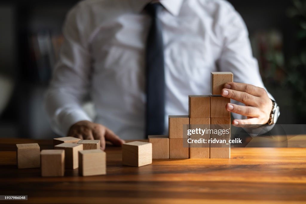 Businessman hand pulling out or placing wood block on the tower and semi-pyramid in modern office. Plan and strategy in business.