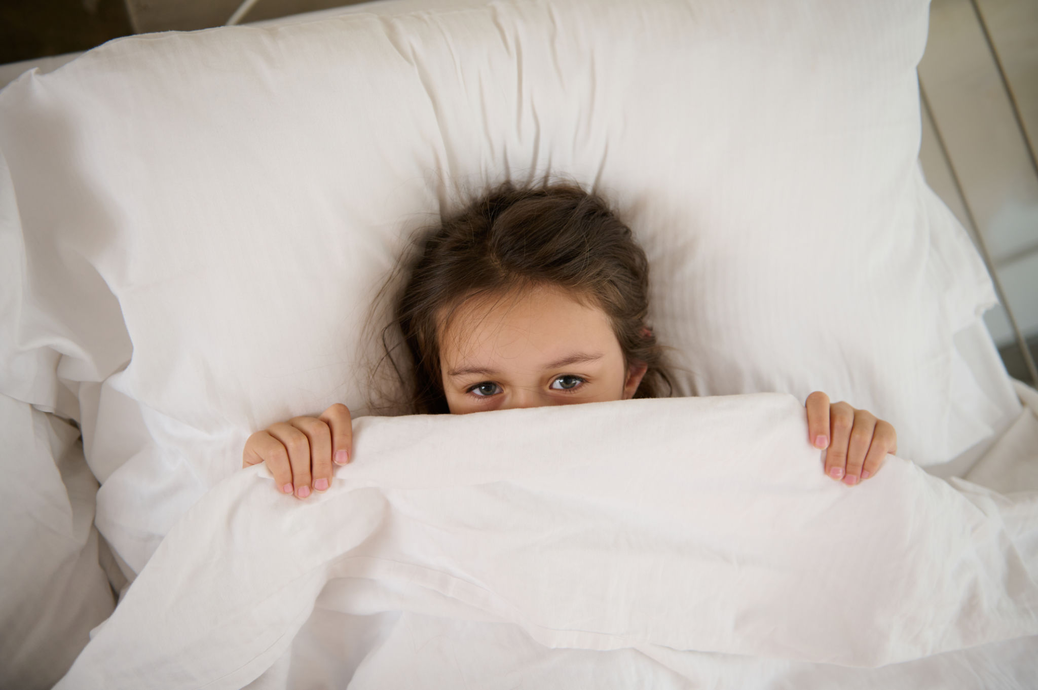 Top view of an adorable child girl lying on the bed, hiding half of her face under white blanket, looking at the camera Top view of an adorable child girl lying on the bed, hiding half of her face under white blanket, looking at the camera