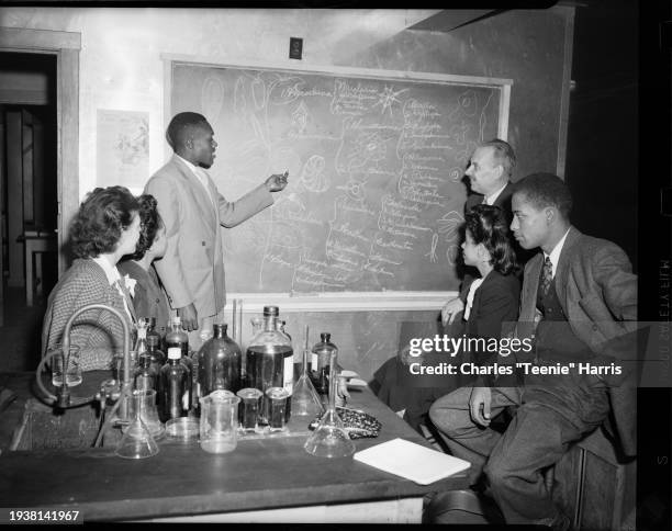 In a classroom, a young man points to a drawing of a spiral shaped micro-organism as others watch, Pittsburgh, Pennsylvania, ca 1940 Various bottles...