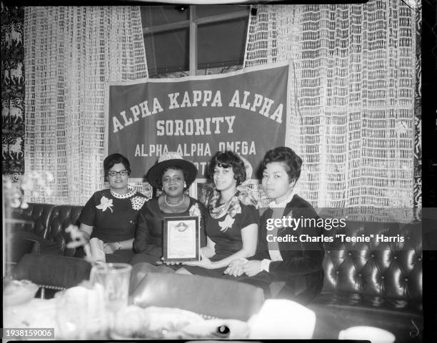 Group portrait of four women, including two holding a plaque, seated on a sofa under a banner that reads 'Alpha Kappa Alpha Sorority, Alpha Kappa...