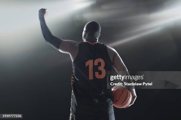 black basketball player with ball raising arm in victorious gesture against dark background - basketball uniform stock pictures, royalty-free photos & images
