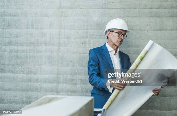 engineer wearing hardhat and examining blueprint near wall - blueprint rolls stock pictures, royalty-free photos & images