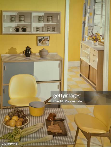 View of an early Sixties kitchen interior decorated in yellow, the scene includes food ingredients and saucepan on a table, dining chairs, wall and...