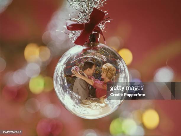 View of a couple embracing in front of a Christmas tree, with the woman holding a wrapped gift, reflected on the surface of a glass bauble...