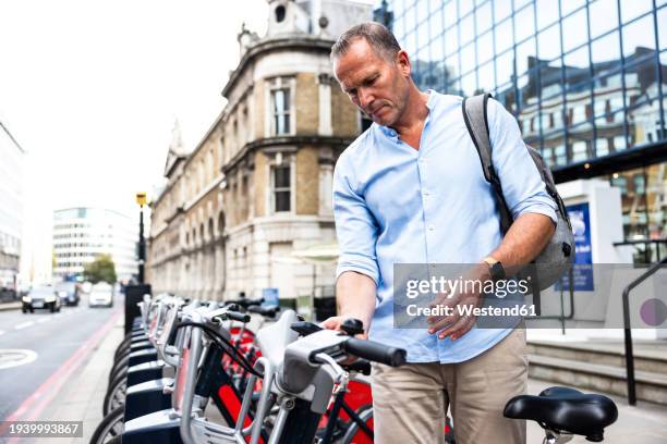 mature businessman renting electric bicycle with smart phone at street - commuter man europe bike stock pictures, royalty-free photos & images