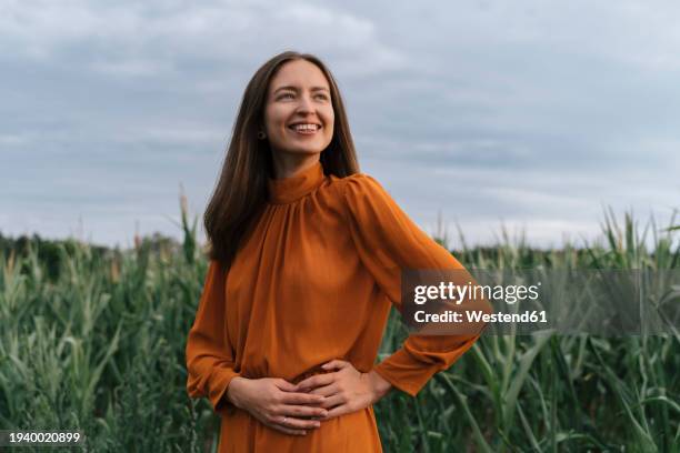 cheerful woman with hands on hip standing by corn crops at field - frau mittleren alters stock-fotos und bilder