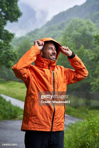 happy man wearing orange jacket and standing in front of mountain - rainwear stock pictures, royalty-free photos & images