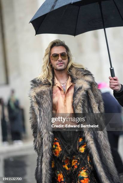 Bill Kaulitz, from the music band Tokio Hotel, wears sunglasses, a brown fluffy oversized faux fur long coat, an orange satin lustrous shirt, black...