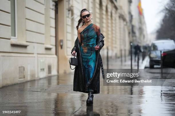 Guest wears sunglasses from Burberry, earrings, tie and dye blue turquoise and brown asymmetric off-shoulder silky long dress, a black leather shiny...