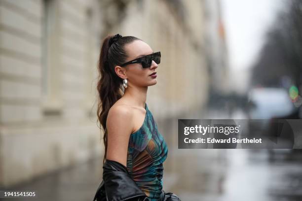 Guest wears sunglasses from Burberry, earrings, tie and dye blue turquoise and brown asymmetric off-shoulder silky long dress, a black leather shiny...