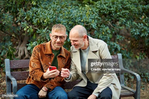 smiling gay men looking at smart phone display together - park bench stock pictures, royalty-free photos & images