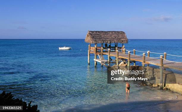 Swimmer enters the water on a small beach and a thatched roof beach cabana with a motorboat moored a few feet offshore, Nassau, Bahamas, 1986. .