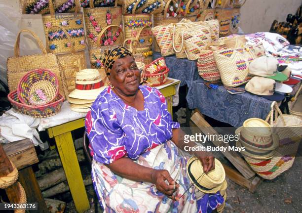 Black woman of African descent sells straw hats, baskets, and other crafts, at an open-air outdoor market in downtown Nassau, Bahamas.
