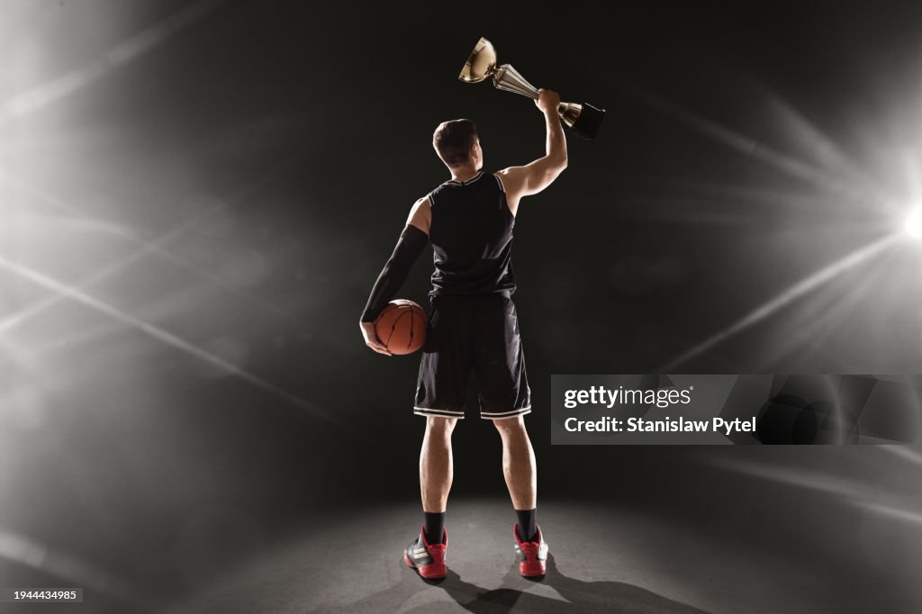 Basketball player standing with ball holding above head a trophy against dark background