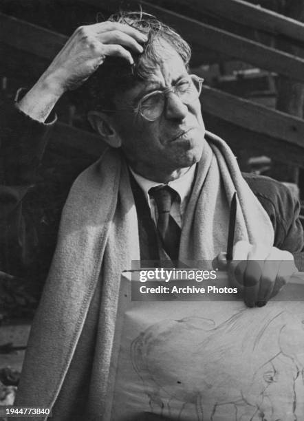 British artist Stanley Spencer scratches his head as he holds a pencil and sketch pad in his other hand, with a scarf around his neck, in his...