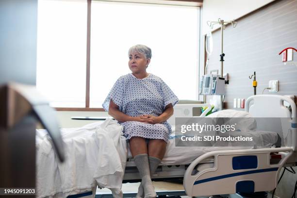 mature female patient sits quietly on the edge of her hospital bed - stretcher stock pictures, royalty-free photos & images