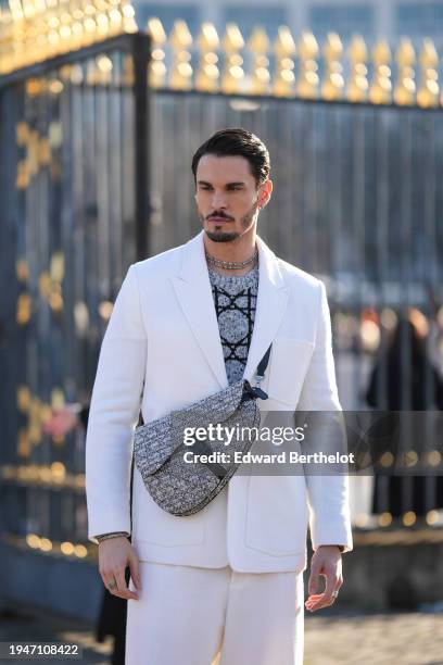 Baptiste Giabiconi wears a necklace, a gray wool Dior pullover, a white blazer jacket, a Dior Saddle tweed bag, white flared pants, loafers shoes,...