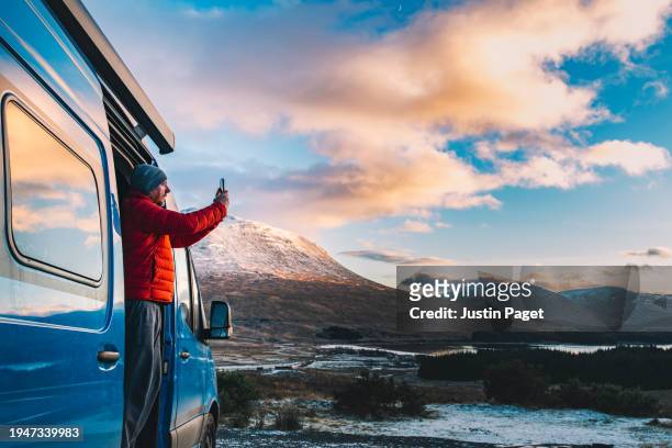 a mature man takes a photo of the view of the stunning snow capped mountains and winter scenery in the scottish highlands from his campervan at sunset - wonderlust stock-fotos und bilder