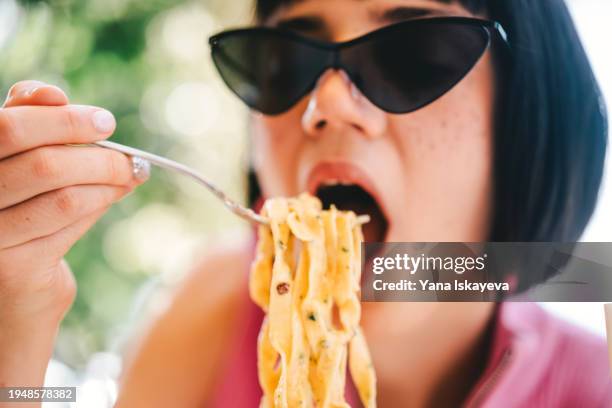 a beautiful young woman in stylish sunglasses eating pasta with pleasure and passion - silver spoon in mouth stock pictures, royalty-free photos & images