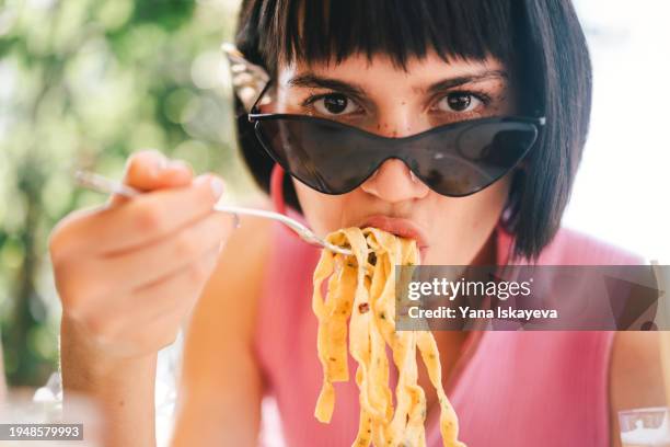 a beautiful young woman in stylish sunglasses eating pasta with pleasure and passion - carbohidrato fotografías e imágenes de stock