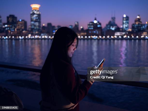 young asian woman using smartphone by the riverside in the city at night - qi yang stock pictures, royalty-free photos & images