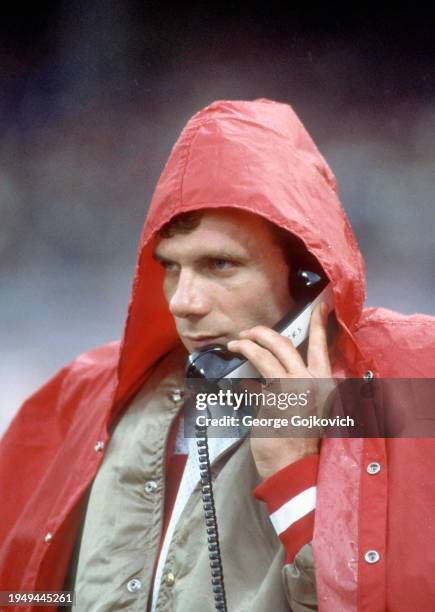 Quarterback Joe Montana of the San Francisco 49ers takes cover under a raincoat hood on the sideline as rain falls as he talks on the phone to...