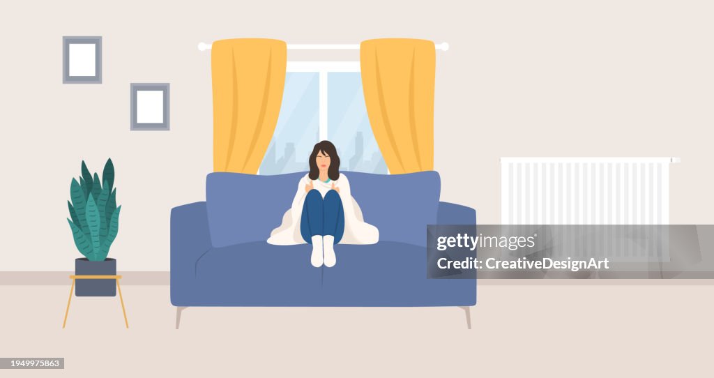 Young Woman Wrapped In Blanket And Sitting On Couch. Living Room Interior With Radiator Heater And Potted Plant. Cold Temperature In The House