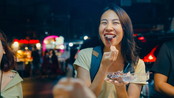 young asian female friends backpacker enjoy eating street food on the street at night market in bangkok, thailand. holiday vacation trip. - chinatown night market stock pictures, royalty-free photos & images