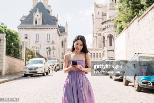 smiling young asian female traveller using smart phone while exploring paris on sunny day in summer. historical french style building in the background. concept of vacation and holiday. - day to the liberation of paris photos et images de collection