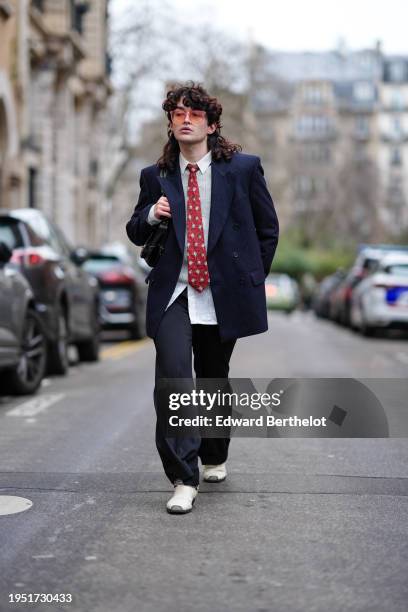 Loic Hornecker wears sunglasses, a white shirt, a red tie, a navy blue blazer jacket, black flared pants, white pointed leather shoes, a bag, outside...