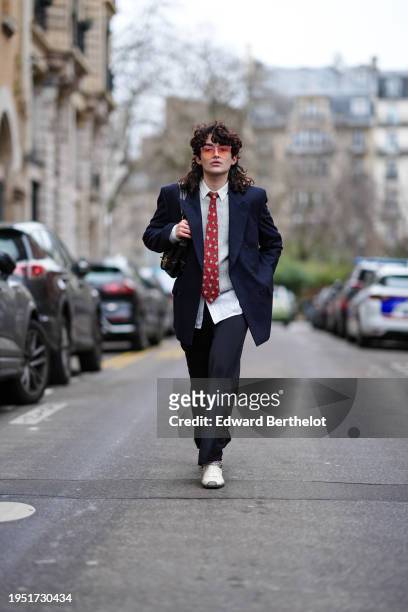 Loic Hornecker wears sunglasses, a white shirt, a red tie, a navy blue blazer jacket, black flared pants, white pointed leather shoes, a bag, outside...