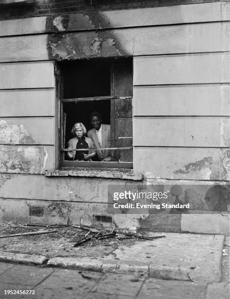 Emanuel Omotola and his wife stand inside their house behind a fire-damaged window following the Notting Hill race riots, London, September 10th 1958.