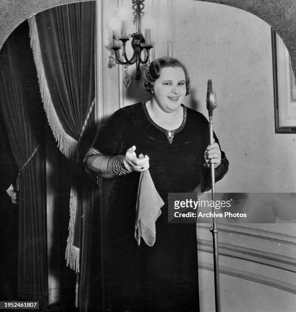 American singer Kate Smith, wearing a dark outfit, smiling as she holds a microphone stand, United States, circa 1935. The image is one-half of a...