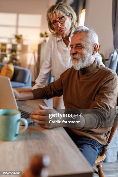 mature man working from home on laptop, his loving wife keeping him company - business mature couple portrait stock pictures, royalty-free photos & images