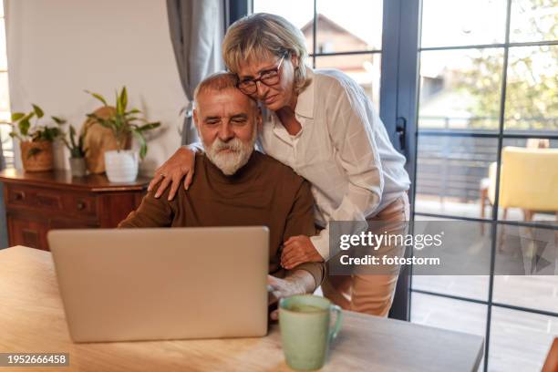 mature man working from home on laptop, his loving wife keeping him company - business mature couple portrait stock pictures, royalty-free photos & images