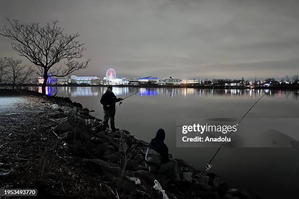 People fishing in the Hackensack River in front of the reflection of the lights of the Ferris wheel of the 'American Dream' shopping center opposite...