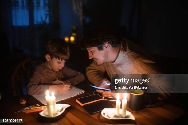 family sitting at home with candles during blackout. child is drawing, man is using solar power bank to charge his smartphone. energy crisis concept. - apagão imagens e fotografias de stock