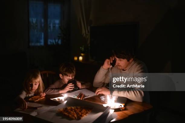familie sitzt zu hause mit kerzen und isst pizza während des stromausfalls. kinder zeichnen, mann ist mann spricht per smartphone. konzept der energiekrise. - kerzenschein stock-fotos und bilder