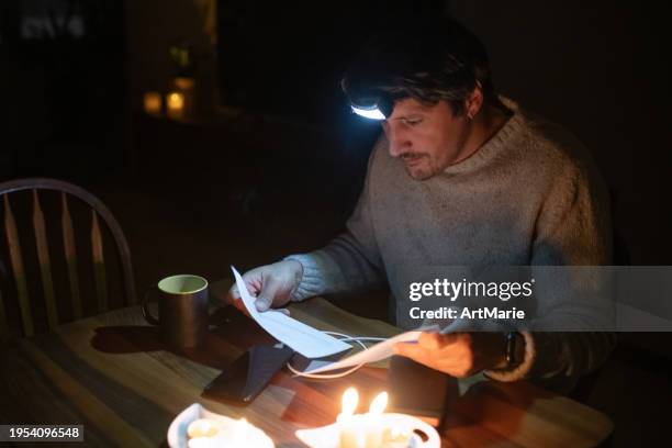 man sitting at home with candles during blackout, examining bills and looking at his smartphone. energy crisis concept. - apagão imagens e fotografias de stock