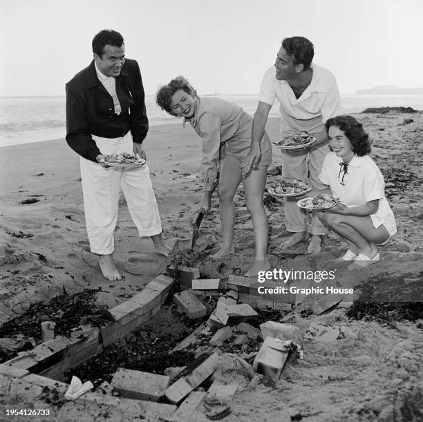 Group comprising two men and two women, around a cooking pit, three holding plates of food as one of the women shovels sand to extinguish the coals...