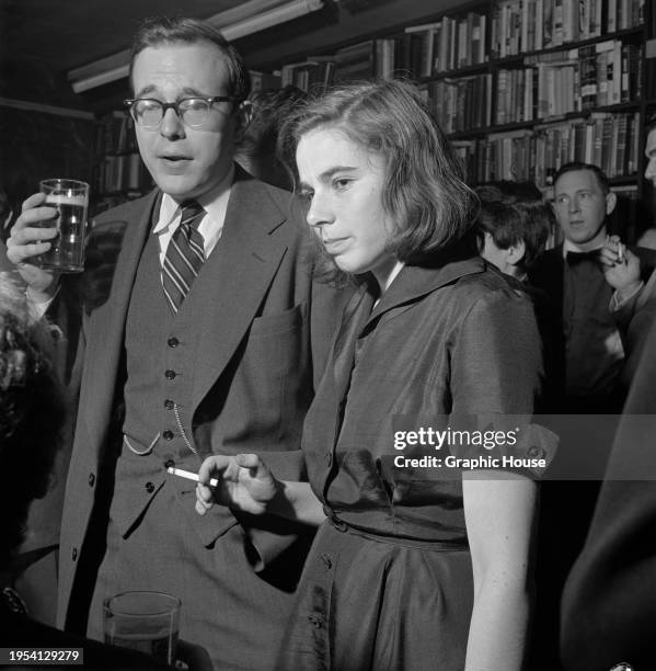 Man wearing a three-piece suit, with a watch chain on his waistcoat, and holding a glass of beer, beside a woman who holds a lit cigarette among...