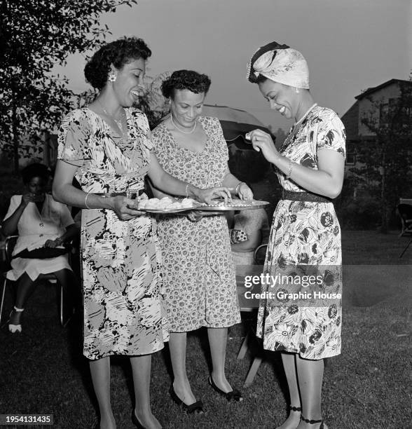 Trinidadian singer and pianist Hazel Scott, wearing a floral pattern short-sleeve dress and a turban, laughing as she holds a piece of food taken...