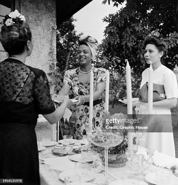 Trinidadian singer and pianist Hazel Scott, wearing a floral pattern short-sleeve dress and a turban, as she takes a plate from a woman at a bridal...