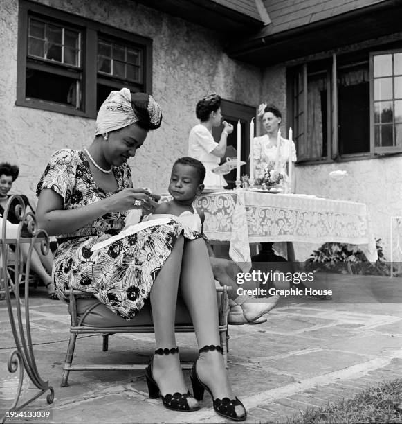 Trinidadian singer and pianist Hazel Scott, wearing a floral pattern short-sleeve dress and a turban, offers food to a small child beside her as she...